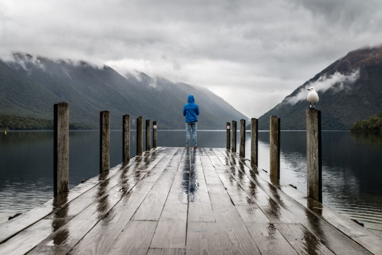 Person in a blue hooded jacket stands at the end of a weathered wooden dock, looking out over a lake.