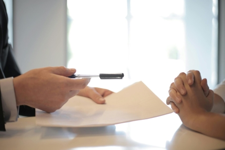 A suited man offers a paper and pen across a table, in a business meeting.