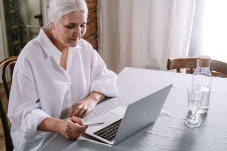 A silver-haired woman is at a table with a laptop.