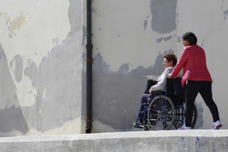 A woman pushes a wheelchair alongside a weathered wall.