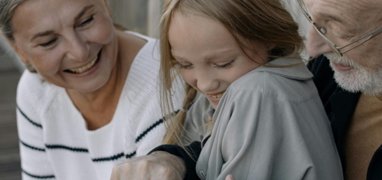 A girl is sitting between her smiling grandparents, looking down with a slight smile herself.