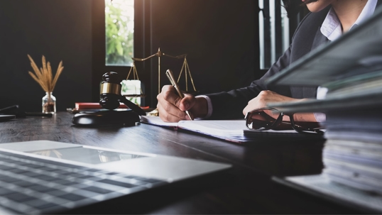 A lawyer in a suit takes notes at a desk with a laptop, gavel, and scales of justice.
